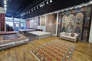 inside-new-store-1- a beige couch on floor with a rug in front of it, stacks of rugs on floor and rugs hung on the wall. black color ceiling and track lights'