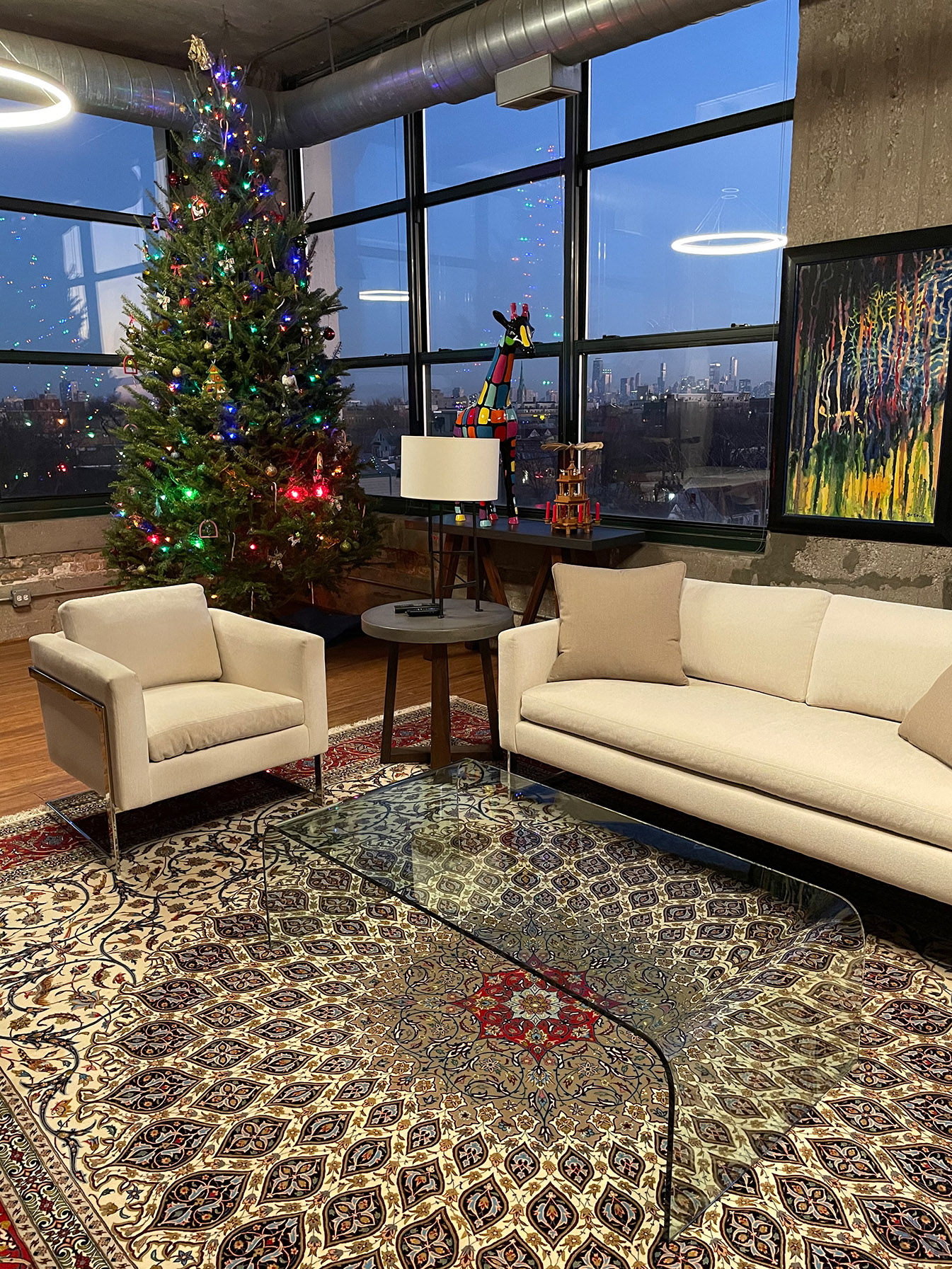 A lining room with a beige Persian rug, glass coffee table and a tall Christmas Tree. High ceiling glas windows