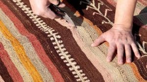 Man is cutting a flat weave rug with a scissor. The rug has brown and white stripes.