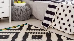 A black and white rug on the floor, a bed with a black and white comforter and white dresser.