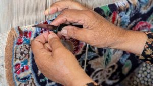 A woman is weaving a Persian rug with knife in her hand trimming the new knot she just made.